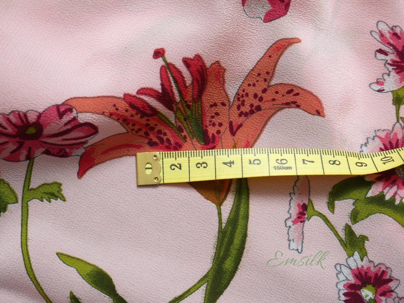 May include: Pink fabric with a floral print featuring a large orange lily with red markings and smaller pink flowers. A yellow measuring tape is placed across the lily, showing measurements in inches.