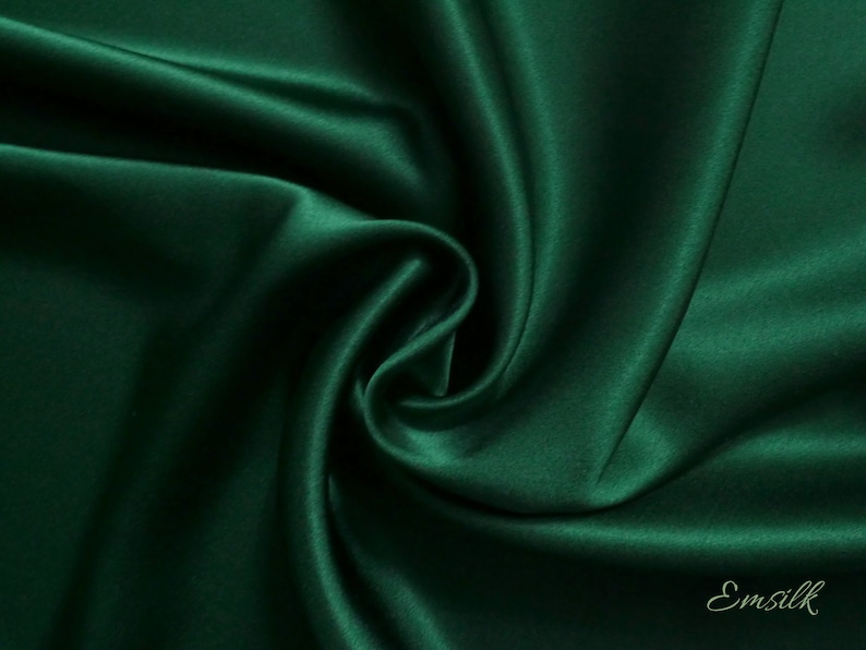 May include: A close-up of a deep green satin fabric, showing its smooth, luxurious texture. The fabric is draped and folded, creating soft curves and folds.