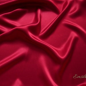May include: A close-up shot of a luxurious, flowing fabric in a deep, rich red hue. The material has a smooth, glossy texture, with soft folds and shadows that create a sense of depth and movement. The word "Emsilk" is in the bottom right corner.