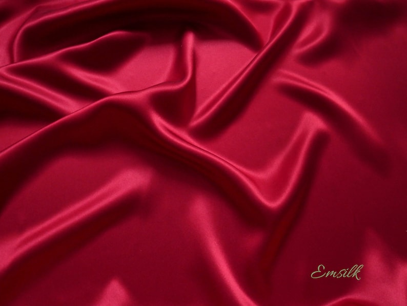 May include: A close-up shot of a luxurious, flowing fabric in a deep, rich red hue. The material has a smooth, glossy texture, with soft folds and shadows that create a sense of depth and movement. The word "Emsilk" is in the bottom right corner.