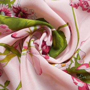 May include: Pink fabric with a floral print featuring pink and green flowers. The fabric is soft and has a subtle texture. The fabric is folded and draped, showing the pattern and texture.