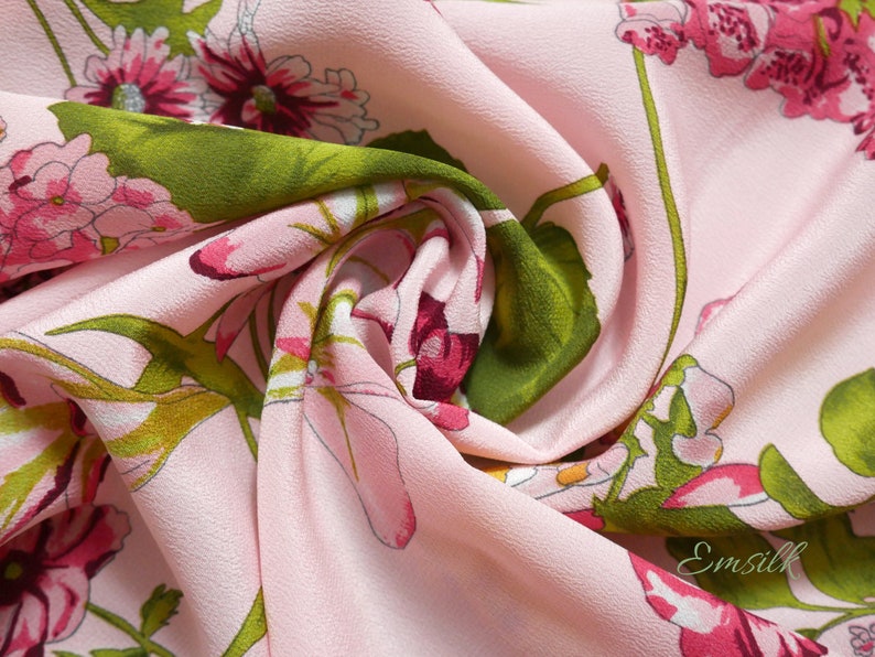 May include: Pink fabric with a floral print featuring pink and green flowers. The fabric is soft and has a subtle texture. The fabric is folded and draped, showing the pattern and texture.