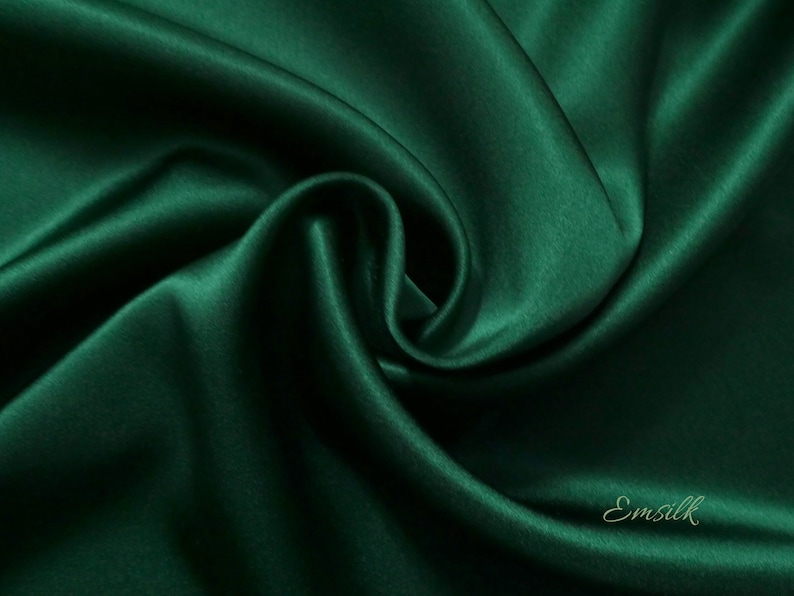 May include: A close-up of a deep green satin fabric with a smooth, silky texture. The fabric is draped and folded, creating soft curves and folds. The fabric is a rich, emerald green color.