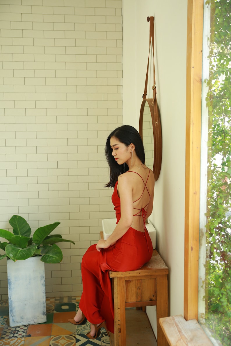 May include: A woman wearing a red dress sits on a wooden stool in a bathroom. The dress has a low back and is tied with a string. The woman is looking down at her feet.