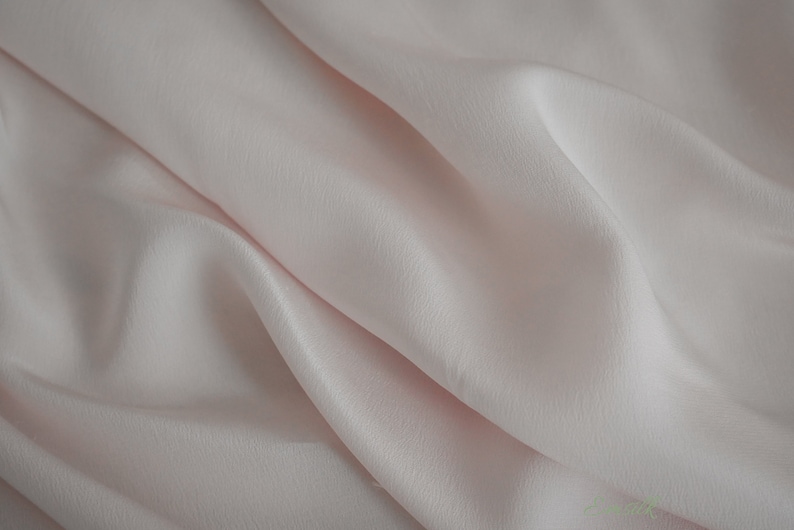 May include: A close-up of a soft, pale pink fabric with a smooth, silky texture. The fabric is draped and folded, creating a gentle, flowing effect.
