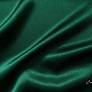May include: A close-up of a deep green satin fabric with soft folds and wrinkles. The fabric is smooth and luxurious, with a subtle sheen. The text "Emsilk" is visible in the bottom right corner.
