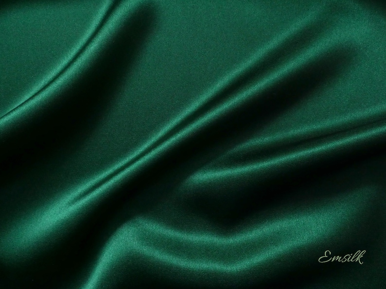 May include: A close-up of a deep green satin fabric with soft folds and wrinkles. The fabric is smooth and luxurious, with a subtle sheen. The text "Emsilk" is visible in the bottom right corner.
