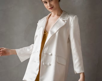 Puede incluir: Una chaqueta blanca de satén con cierre de doble botonadura y un vestido de satén dorado debajo. La chaqueta tiene dos botones grandes y un bolsillo en el lado izquierdo. La modelo lleva la chaqueta con el cuello levantado.