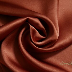 May include: Close-up of a luxurious, flowing fabric in a rich, warm terracotta color. The material has a smooth, satiny texture with subtle highlights and shadows, creating a sense of depth. The word "Emsilk" is printed in a cursive font.