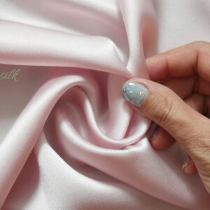 May include: A close-up of a soft, pink satin fabric. The fabric is draped and folded, showing its smooth texture. The text "Emsilk" is visible in the upper left corner of the image.