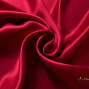 May include: Close-up of a swirl of deep red satin fabric. The fabric has a smooth, lustrous texture with highlights and shadows that emphasize the folds and curves. The word "Emsilk" is written in a cursive font in the bottom right corner.
