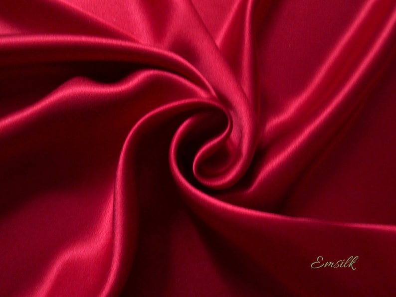 May include: Close-up of a swirl of deep red satin fabric. The fabric has a smooth, lustrous texture with highlights and shadows that emphasize the folds and curves. The word "Emsilk" is written in a cursive font in the bottom right corner.