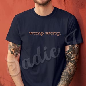 Può includere: T-shirt blu navy girocollo con la scritta "womp womp." in arancione. La maglietta a maniche corte è di colore unito. Il testo è centrato sul petto.