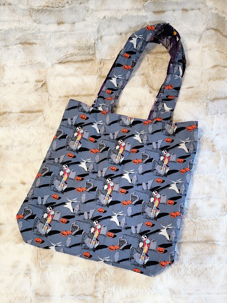 May include: A blue tote bag featuring a repeated pattern of Jack Skellington and Zero from The Nightmare Before Christmas. The design includes pumpkins, tombstones, and a graveyard setting.