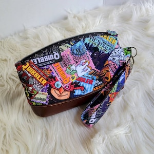 May include: A colorful zippered pouch with a brown leather bottom and a matching wrist strap. The pouch is decorated with a print of newspaper clippings and illustrations, including a unicorn, a wizard, and the words "The Quibbler".