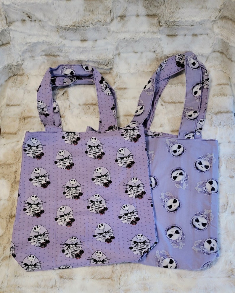 May include: Two purple tote bags with a white polka dot pattern. One bag features a repeating pattern of Jack Skellington from The Nightmare Before Christmas with the text "The Skeleton King". The other bag features a repeating pattern of Jack Skellington's face.