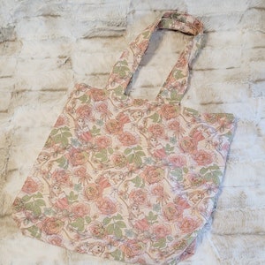 May include: A beige tote bag with a floral pattern featuring pink roses and green leaves. The bag has a single strap and is made of a soft, canvas-like material.
