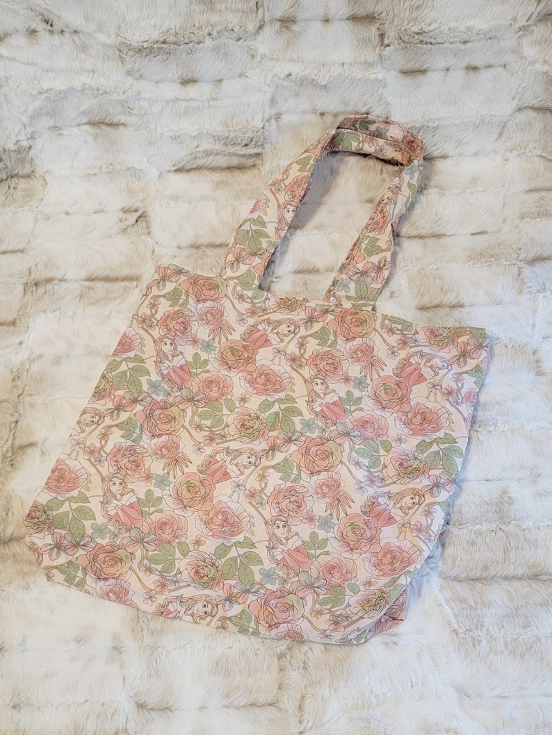 May include: A beige tote bag with a floral pattern featuring pink roses and green leaves. The bag has a single strap and is made of a soft, canvas-like material.