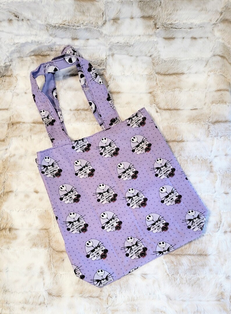 May include: Purple tote bag with a white polka dot pattern and a repeated image of Jack Skellington from The Nightmare Before Christmas. The image includes the text "The Skeleton King".