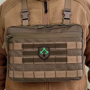 以下が含まれることがあります： ジッパー留めと複数のMOLLEウェビングの列を持つ茶色の戦術的なチェストリグ。幾何学的なデザインの緑色のパッチが前面に取り付けられています。