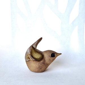May include: A small, hand-sculpted ceramic bird figurine in a warm, earthy tan colour. The bird features a smooth, rounded body, a pointed beak, a dark, circular eye, and a raised tail. The wing area is highlighted with a subtle, mottled yellow-brown glaze.