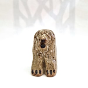 May include: A small, hand-sculpted ceramic figurine of a dog in a seated position. The piece features a speckled, light brown glaze with darker brown accents on the face, ears, and paws, showcasing a textured, earthy finish throughout the body.