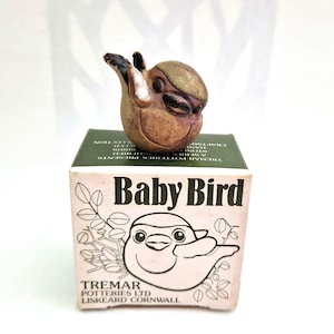 May include: A small brown ceramic bird figurine with a rounded body and dark markings, perched atop a square cardboard box. The box features the text Baby Bird, Tremar Potteries Ltd, Liskeard Cornwall, and a line drawing of a bird on a branch.