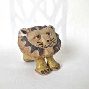 May include: A small ceramic figurine of a lion with a tan body and a dark brown mane. The lion has painted black facial features including eyes, whiskers, and a mouth. The figure stands on two front legs and has a textured, hand-crafted appearance.