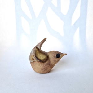 May include: A small, hand-sculpted ceramic bird figurine with a speckled tan and brown finish. The bird features a pointed beak, a dark circular eye, and a raised tail, with a distinct textured wing area highlighted in a darker, muted olive-brown tone.