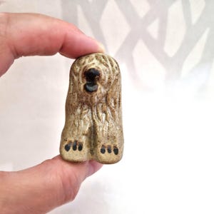 May include: A small, hand-sculpted ceramic figurine of a shaggy dog with a speckled beige and brown glaze. The dog has a textured coat, a dark brown nose, a dark brown open mouth, and dark brown painted paw details on its front feet.