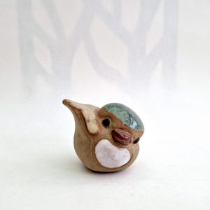 May include: A small ceramic bird ornament. It has a brown body, a teal head, and a white chest. The bird has a small beak and dark eyes. The ornament is set against a white backdrop.