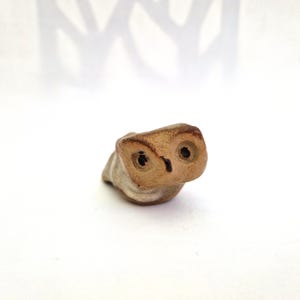 May include: A small, hand-sculpted ceramic owl figurine with a light tan and brown speckled finish. The owl features two dark, circular indentations for eyes and a small, pointed beak, with a smooth, rounded body shape and a flat base.