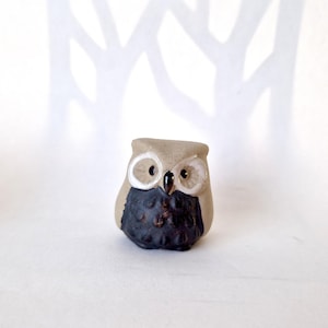 May include: A small ceramic owl figurine featuring a light beige upper body and a dark brown, textured lower body. The owl has two prominent white circular rings around its black eyes and a small, dark, pointed beak centred on its face.