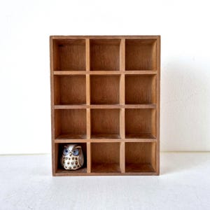 May include: A rectangular wooden display shelf featuring twelve square compartments arranged in a four-by-three grid. A small, ceramic owl figurine with brown and white markings and blue eyes sits inside the bottom-left compartment.