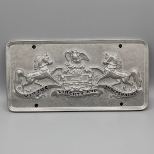 May include: A rectangular, silver-colored metal license plate with embossed details. The design features two rearing horses flanking a central crest with an eagle and the words "VIRTUE LIBERTY AND INDEPENDENCE". Four holes are visible for mounting.