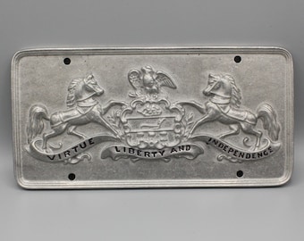 May include: A rectangular, silver-coloured metal license plate with embossed details. The design features two rearing horses flanking a central crest with an eagle and the words "VIRTUE LIBERTY AND INDEPENDENCE". Four holes are visible for mounting.