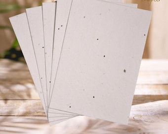 May include: A stack of off-white rectangular seed paper sheets with small dark flecks. The paper is fanned out, revealing the edges of each sheet. The Green Chapter logo is visible in the upper right corner. The paper rests on a wooden surface.
