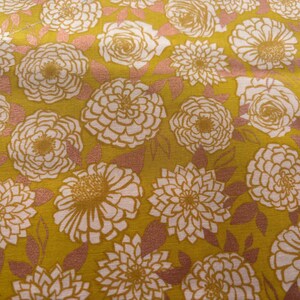 May include: A close-up of a fabric with a repeating floral pattern. The design features large white and gold flowers, including roses and dahlias, set against a mustard yellow background. Pinkish-brown leaves fill the spaces between the flowers.