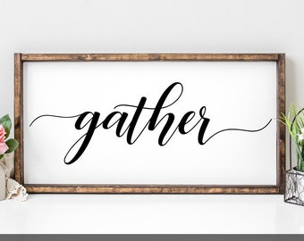 Op de afbeelding: Een rechthoekig houten bord met het woord "gather" in elegant zwart schrift. Het bord heeft een witte achtergrond en een donkerbruine houten lijst. Het woord is geschreven in een vloeiend, cursief lettertype, ideaal voor woondecoratie.