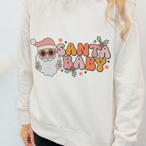 May include: White sweatshirt with a retro-style graphic of Santa Claus wearing sunglasses and the text "Santa Baby" in colorful letters.