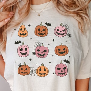 以下が含まれることがあります： ピンク、オレンジ、ブラックのハロウィンプンプキンデザインの白いTシャツ。パンプキンはそれぞれ異なる表情をしていて、コウモリ、星、クモの巣で囲まれています。