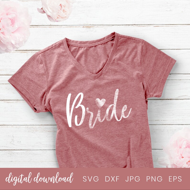 May include: A pink t-shirt with the word "Bride" in white script with a small heart next to it. The shirt is on a white wooden background.