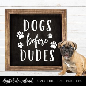 以下が含まれることがあります： 白い文字で「Dogs before Dudes」と書かれた黒いチョークボードの看板。文字の周りに肉球の跡が描かれている。看板の前に、茶色と白の小さな子犬が座っている。