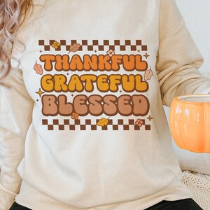 Peut inclure: Un sweat-shirt beige avec le texte "Thankful Grateful Blessed" en lettres brunes, orange et jaunes avec un motif &agrave; carreaux et des feuilles d'automne.