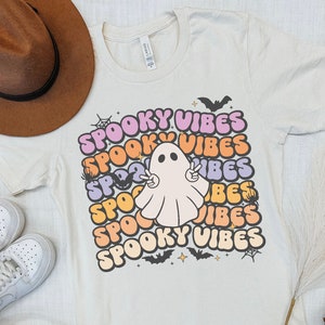 May include: A white t-shirt with a colorful graphic design featuring the text "Spooky Vibes" and a cartoon ghost with outstretched arms. The ghost is in a light pink color.
