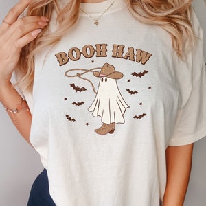 May include: A white t-shirt with the text "Booh Haw" printed on it. The text is in a brown font with a cowboy hat and ghost graphic. The ghost is wearing a cowboy hat and holding a lasso.