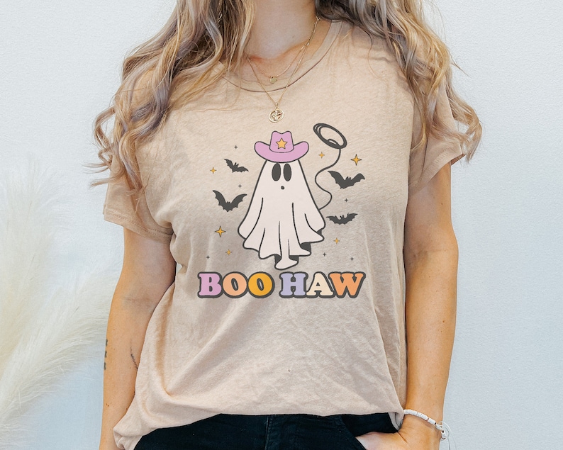 Puede incluir: Una camiseta beige con un fantasma de dibujos animados que lleva un sombrero de vaquero rosa y el texto "BOO HAW" en letras de colores.