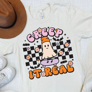May include: A white t-shirt with a pink and orange graphic of a ghost skateboarding. The graphic says "Creep It Real" with a checkered background and pink flowers.