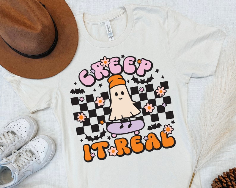 May include: A white t-shirt with a pink and orange graphic of a ghost skateboarding. The graphic says "Creep It Real" with a checkered background and pink flowers.