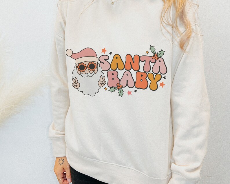 May include: White sweatshirt with a retro-style graphic of Santa Claus wearing sunglasses and the text "Santa Baby" in colorful letters.